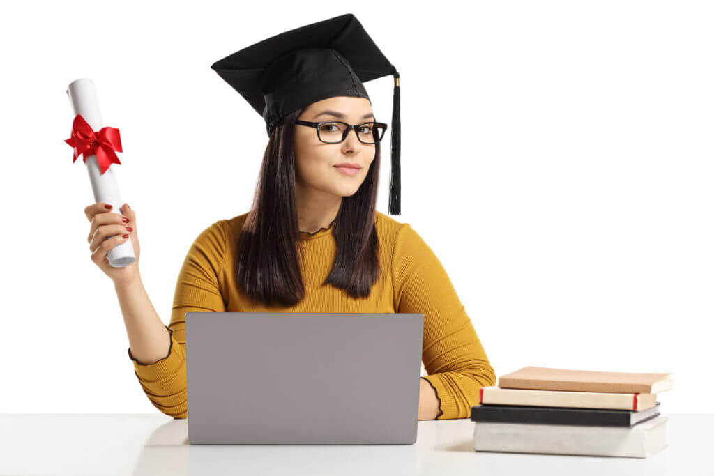 Young woman with a graduation hat and diploma, sitting in front of a laptop. 